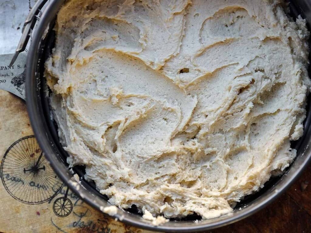 A close-up of a round baking tin filled with thick, creamy cake batter, ready to be baked. The tin sits on a rustic surface with vintage-style designs and text partially visible underneath.